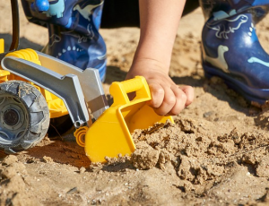 ein Kind in Gummistiefeln spielt mit einem Spielzeugbagger im Sandkasten
