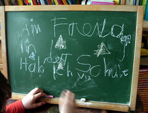 eine Kreidetafel, auf der in Kinderschrift einige Worte gekritzelt sind, man kann lesen: Am Freitag in der Schule