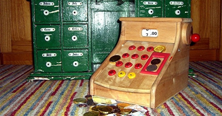 eine Spielzeug-Kasse mit Spielgeld vor einem altmodischen Kaufmannsladen